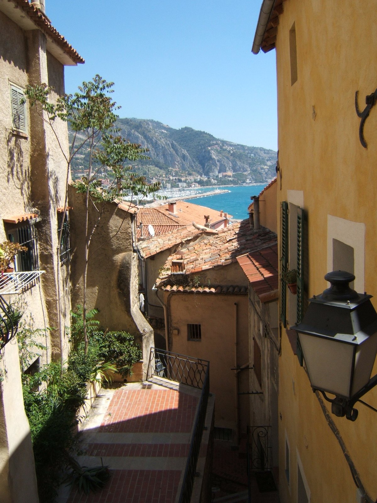 Smal, fargerik gate i gamle Menton med varme fasader, skodder og utsyn videre mot havnen i bakgrunnen.